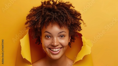 A happy young woman with curly hair is posing with her head in a hole in a paper, looking through a
