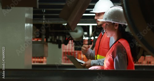 Male and female rail engineers, wearing orange safety vests and helmets, inspecting the underside of an electric train system in an industrial and engineering setting.