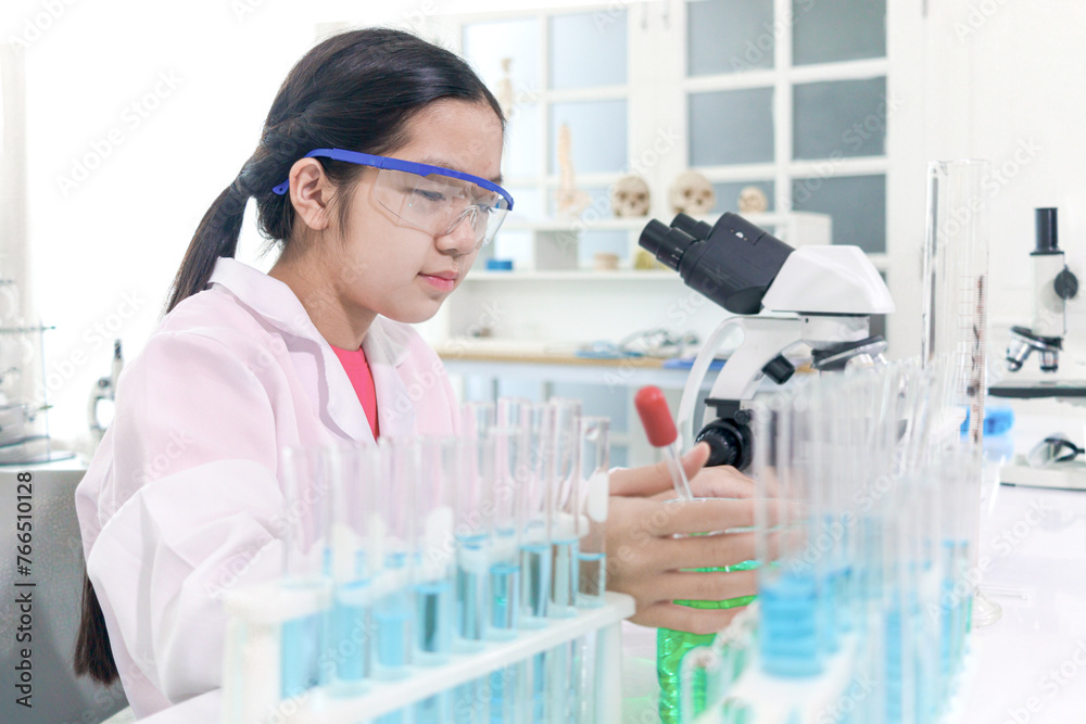 Cute young scientist schoolgirl wearing lab coat and safety glasses, doing science experiments. Student girl child using lab equipment to study chemistry in laboratory. Kid learning science education.