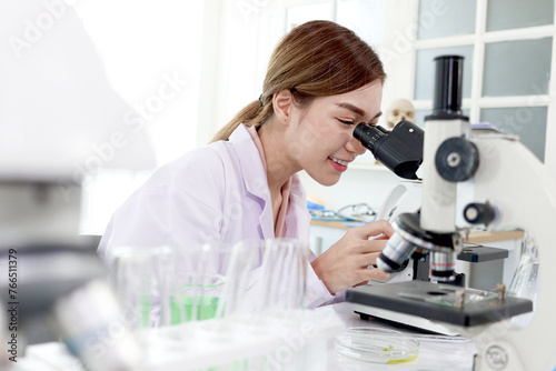 Beautiful Asian female scientist in lab coats does science experiments, happy researcher woman looks through microscope, uses equipment for research. Woman works in science and bio medical field.