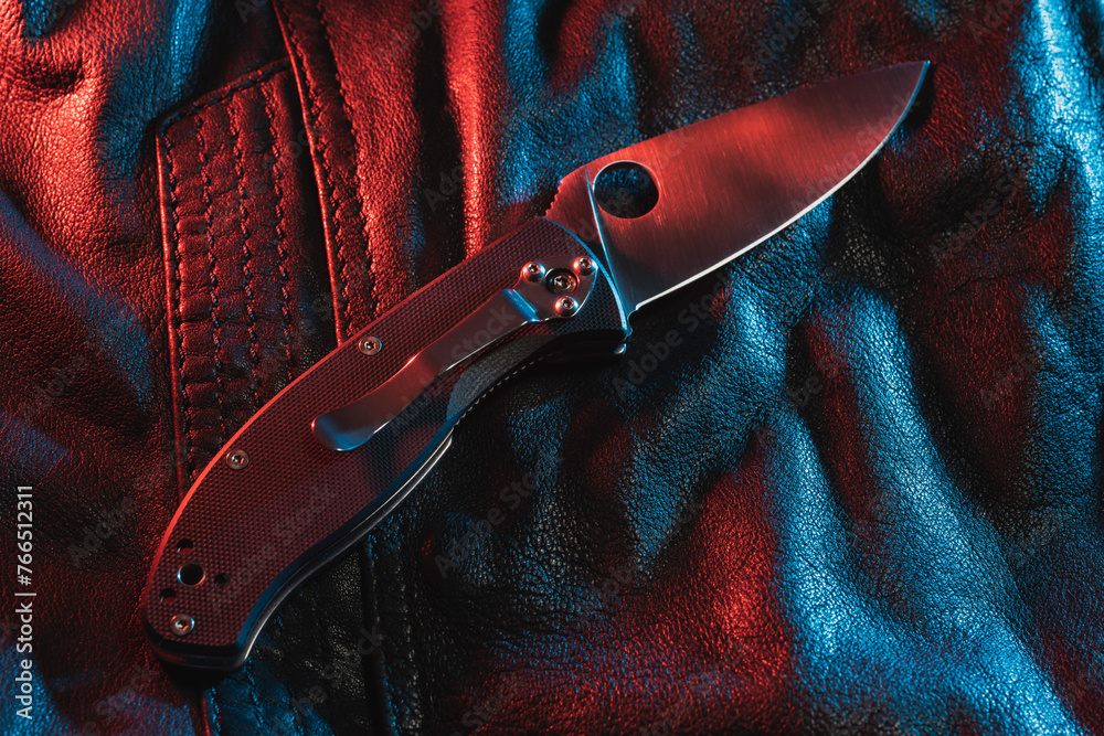 A folding knife lies on a leather jacket, multi-colored lighting. - obrazy, fototapety, plakaty 