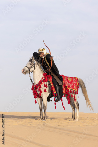 Saudi Man with his white stallion in a desert, shooting an arrow