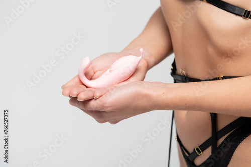 Close-up view of slim caucasian woman wearing black lingerie and holding small pink vibrator next to her belly against gray background. Soft focus. Adult sex toys theme.