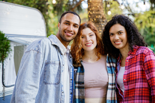 A group of friends are relaxing in nature. Two girls and a guy of different races are looking at the camera, smiling, having fun on the background of a travel trailer.