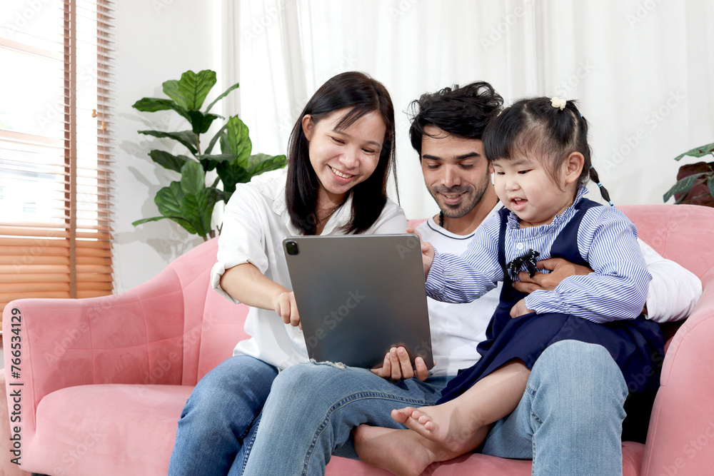 Fototapeta premium Happy family in living room, Chubby little girl daughter with her mother and father using tablet in living room. Kid spending online together with parents at home. Child, mom and dad in house.