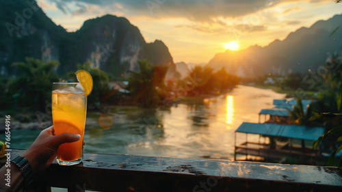 Hand holding a refreshing drink against a scenic sunset backdrop over a tranquil river surrounded by mountains. photo