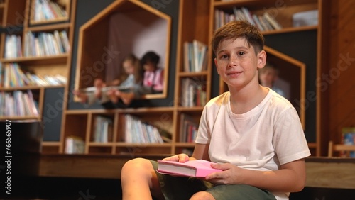 Caucasian boy looking at camera while group of smart students sitting at library. Child studying  learning  reading from novel or textbook while children talking  chitchat about education. Erudition.