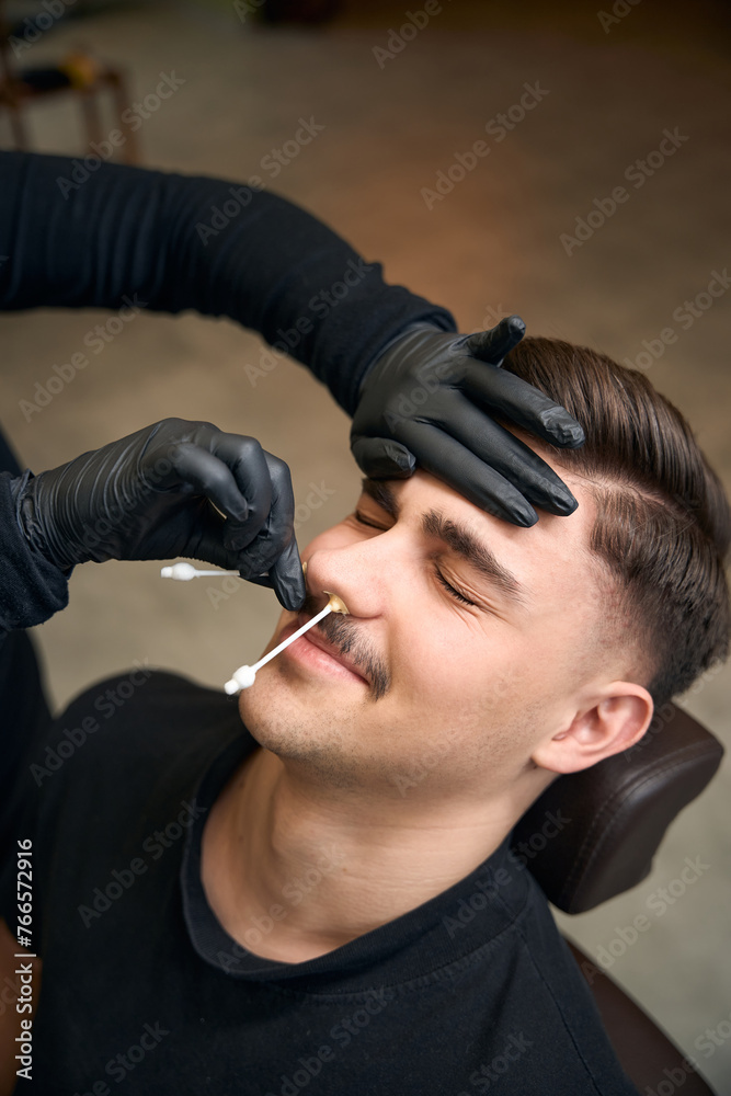 Unrecognizable master barber removes hair from nose of man with wax