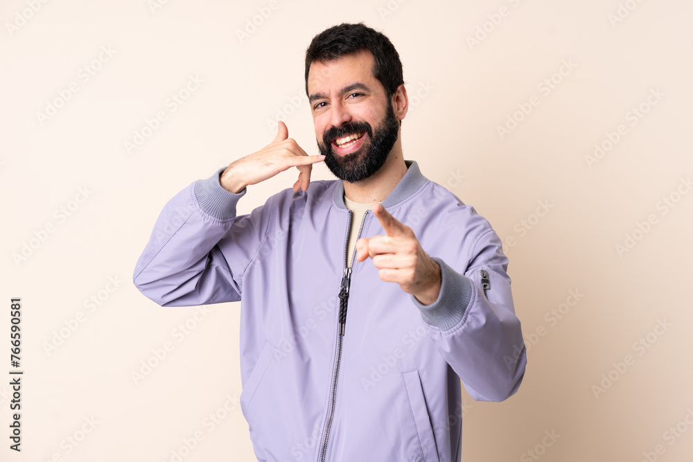Caucasian man with beard wearing a jacket over isolated background making phone gesture and pointing front Caucasian man with beard wearing a jacket over isolated background making phone gesture and pointing front