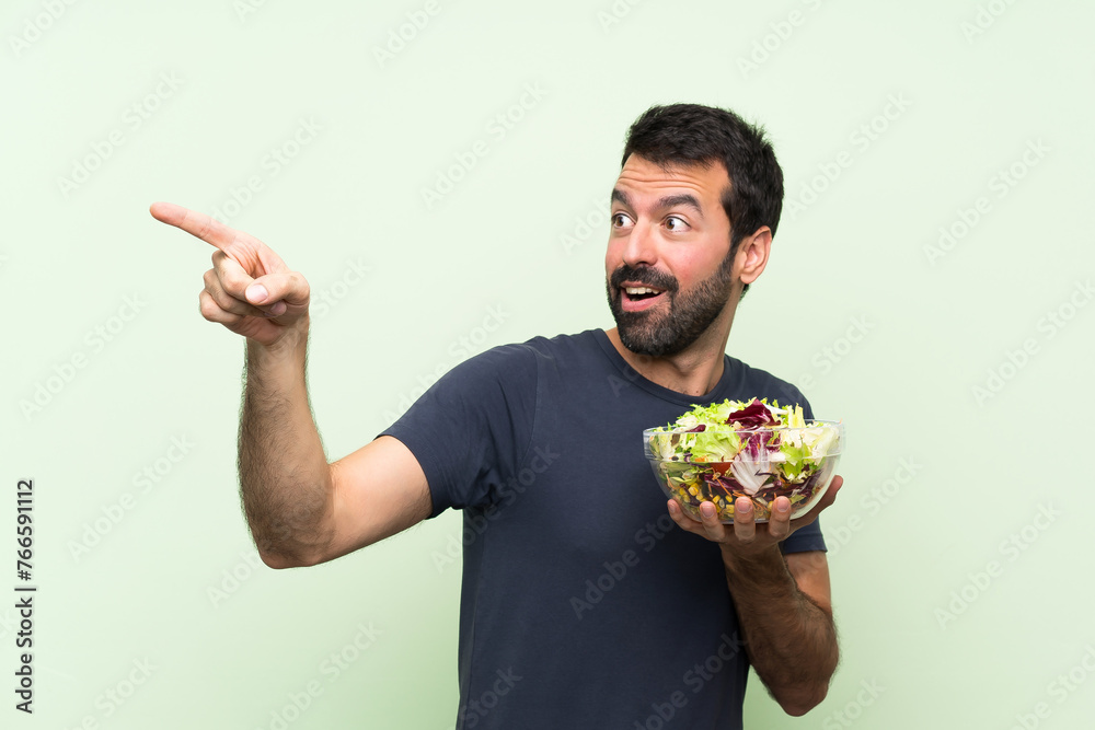 Young handsome man with salad over isolated green wall pointing away Young handsome man with salad over isolated green wall pointing away