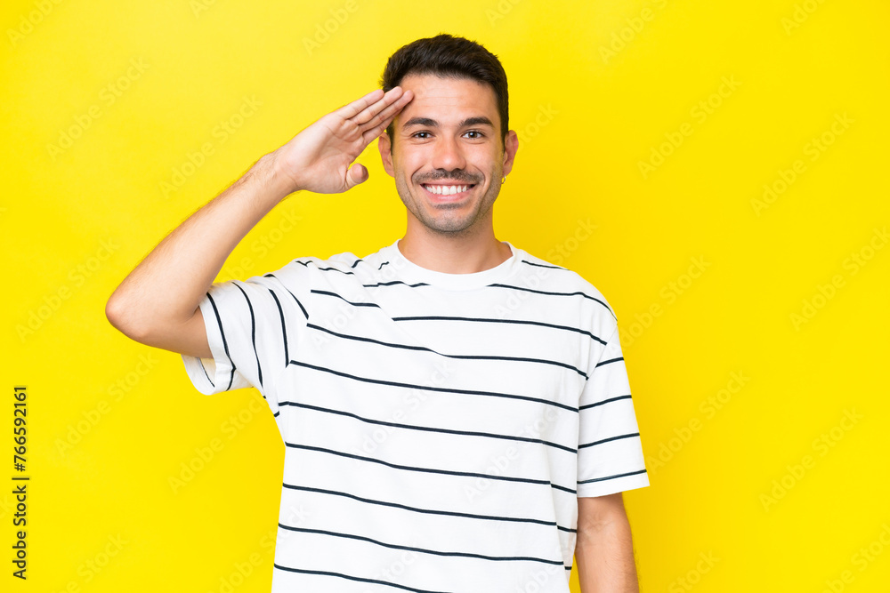 Young handsome man over isolated yellow background saluting with hand with happy expression Young handsome man over isolated yellow background saluting with hand with happy expression