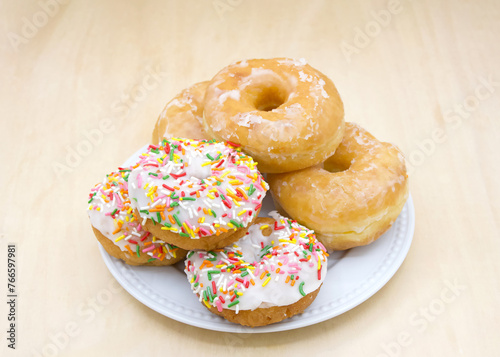 White porcelain plate with glazed and frosted cake donuts with sprinkles on a light wood table.