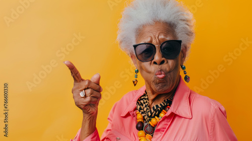 A spirited elderly woman with sunglasses pointing with panache against an orange backdrop.