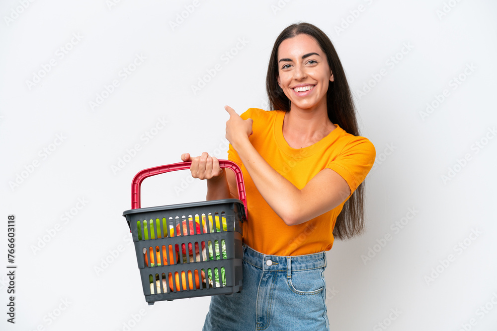Young caucasian woman holding a shopping basket full of food isolated on white background pointing back Young caucasian woman holding a shopping basket full of food isolated on white background pointing back