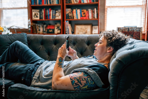 LGBTQ+ person lounging on a sofa, comfortably using a smartphone, depicting everyday life and modern connectivity.

 photo