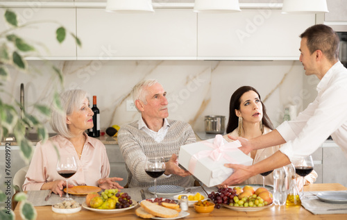 Caring son-in-law surprising father-in-law with present during cheerful family feast while mother-in-law and wife watching with smiles, sitting at set table in well-appointed light kitchen