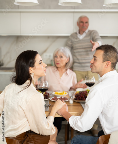 Young man and woman flirting sitting around table together with old family members who are watching them with objection