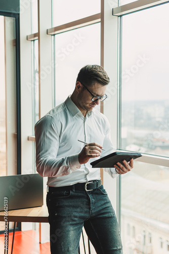 A handsome man in glasses and a shirt stands in the office at the table and works on a tablet. A modern workspace with office space