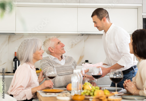 Birthday senior man accepts valuable gift greetings from adult son during family feast. Mature parents celebrate family holiday together with their son and daughter-in-law