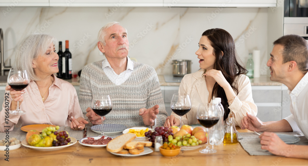 Cozy dinner scene while elderly parents enjoying wine and meal with adult daughter and son-in-law in well-lit home kitchen..