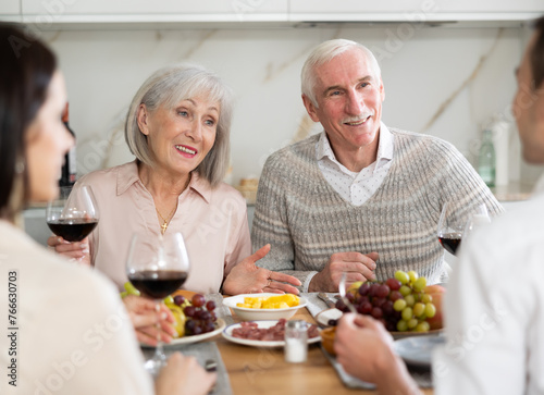 Happy senior parents talking with young couple while drinking wine sitting around kitchen table