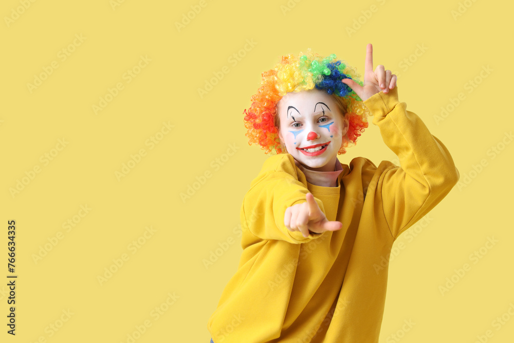 Funny little girl in clown wig showing loser gesture on yellow background. April Fools' Day celebration Funny little girl in clown wig showing loser gesture on yellow background. April Fools' Day celebration