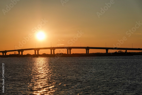 Sunset over a long bridge with an orange sky and reflection on the bay water