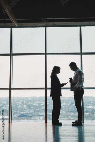 A man and a woman are standing in the office against the background of a large panoramic window. Modern spacious workspace with employees. Business partners