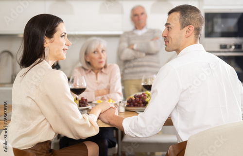 Couple sharing close moment at dining table, holding hands and locking eyes, while perplexed parents watching in background at family home