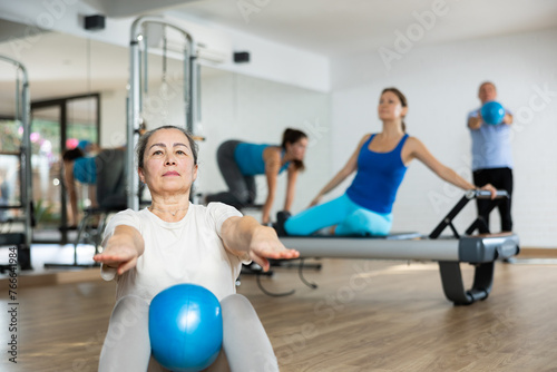 Slender young woman practicing pilates with ball in fitness hall during workout session. Persons doing pilates with trainer