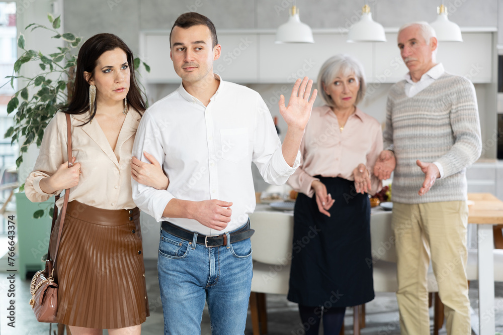 Fototapeta premium Young adult man gesturing decisively to troubled perplexed elderly parents, seeking to put stop to family argument, while leaving home with girlfriend..