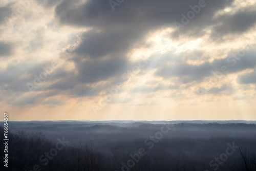 An early morning scenic view of rolling forest in southern Indiana in the early spring. The sky is mostly cloudy with rays of sunshine. You can see the horizon. A nice background image. 