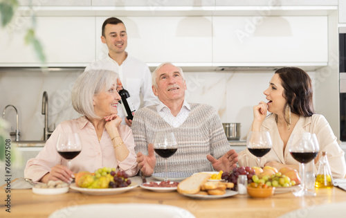 Mature couple are sitting at table, chatting sweetly, discussing and telling news, sharing plans with adult children. Senior woman and man celebrate housewarming party with middle-aged guests spouses