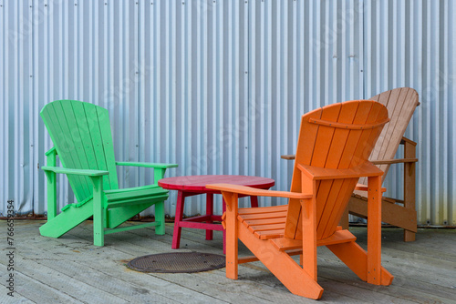 Three colorful Adirondack high-back chairs  orange  green  and pink in color  on a wooden deck with a grey metal corrugated wall. A small red round plastic table is in the center of the seating.  