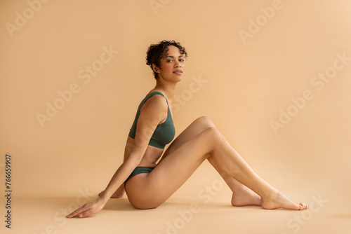 Beautiful black woman in green underwear, sitting on floor and posing at camera over beige studio background, full length, free space