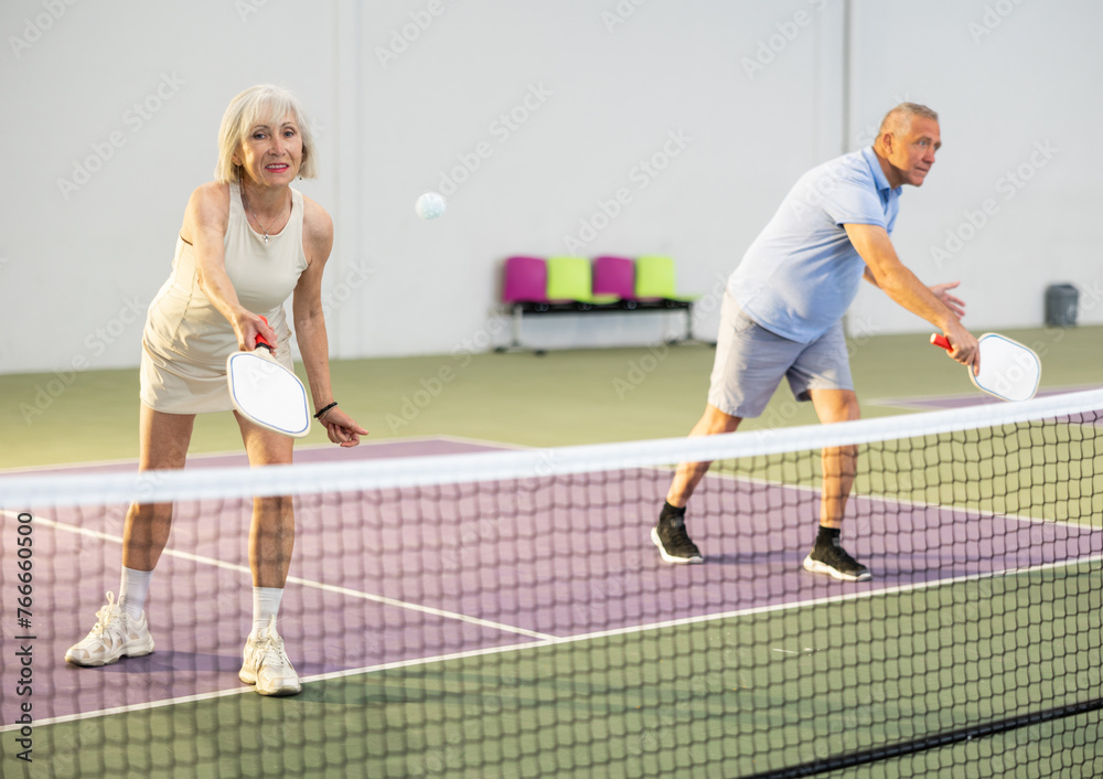 Obraz premium Focused positive elderly woman playing friendly pickleball match in team with male partner on indoors court. Concept of concentration in competition