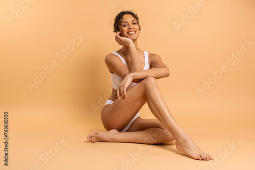Love Yourself. Lady in underwear sitting on floor and smiling at camera over beige background, woman enjoying her natural beauty