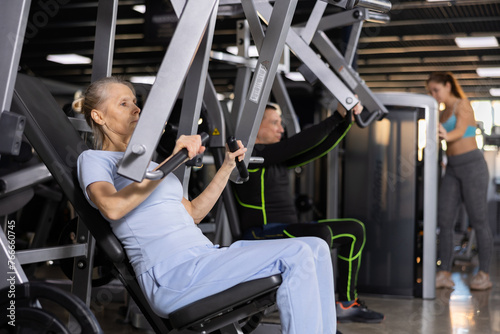 Concentrated aged woman leading healthy active lifestyle doing strength training in gym, performing chest press in exercise machine