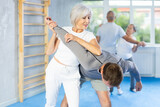 Elderly woman twists the arm of attacking man with painful hold in gym. Self-defense lesson