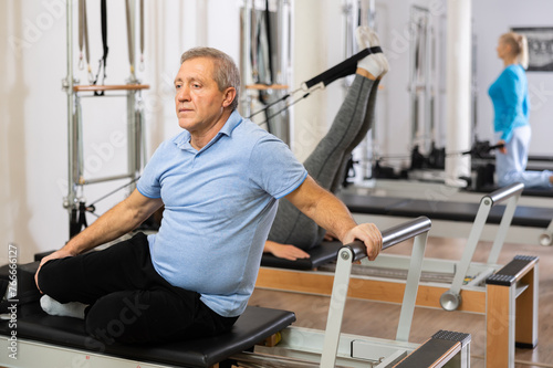 Senior male in activewear performing mermaid exercise on reformer Pilates machine for improving and maintaining mobility in rehabilitation center © JackF