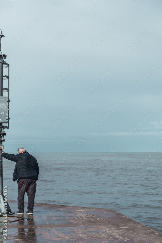 Obraz premium Senior man leaning on the headlight of the pier in a Riviera Romagnola beach location. Dramatic sky in a winter afternoon. Winter sea.