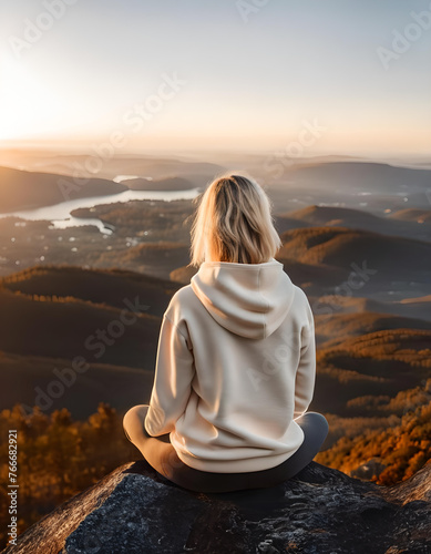 Women overlooks beautiful mountain vista at sunrise. Tourist views river from cliff with short blonde hair and blank hoodie. 