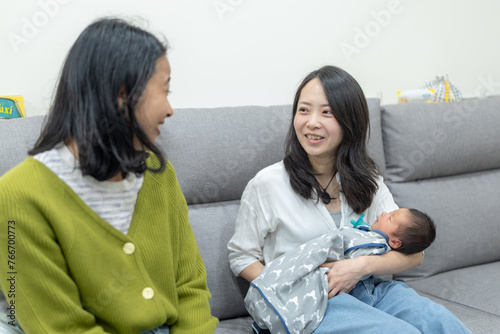 30代の台湾人女性の母親が生後8日の新生児を持って横の20代のアジア人女性の友達と室内のソファに座って話している The mother of a Taiwanese woman in her 30s is sitting on an indoor sofa with an Asian woman in her 20s next to her with an 8 days old newborn b