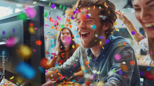 Colleagues celebrating with confetti at work - Joyful office celebration with laughing colleagues and colorful confetti flying around a computer desk