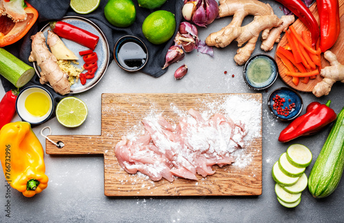 Food and cooking background. Gray table with chopped chicken slices with starch. Paprika, zucchini, vegetables, spices and ingredients for cooking Asian dishes with ginger, garlic, soy sauce, top view