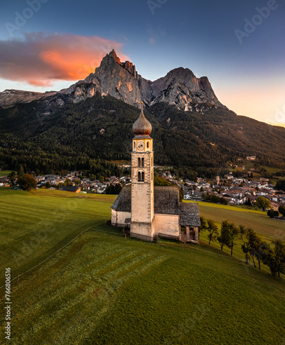 Seis am Schlern, Italy - Aerial view of St. Valentin Church and famous Mount Sciliar mountain at background at sunset with golden and blue sky and warm sunlight at South Tyrol on a summer afternoon
