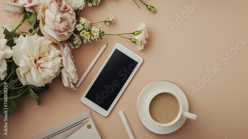 Top view of a tablet, notepad, and smartphone next to a milk-filled coffee cup and flower bouquet on a light brown background.
