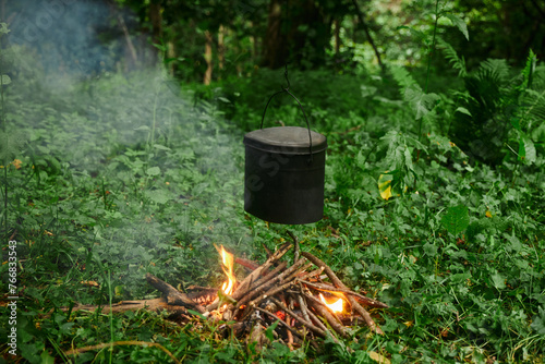 Black cooking pot hangs over a crackling campfire surrounded by dense foliage