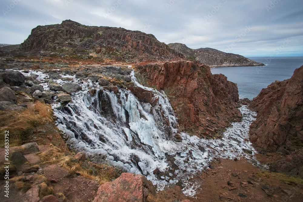 Waterfall tumbles over rocks into the ocean under a cloudy sky as evening approaches. Teriberka, Russia
