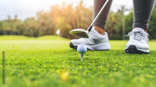 A person is getting ready to hit a golf ball with a golf club. The ball is on a tee and the person is wearing white shoes. The background shows a golf course 